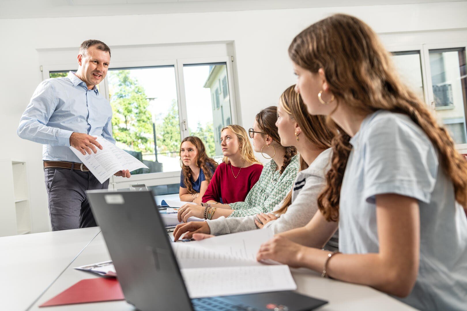 Ein Mann steht vorne an einem Konferenztisch, hält Papiere in der Hand und spricht zu fünf Frauen, die aufmerksam zuhören. Laptops und Dokumente liegen auf dem Tisch, große Fenster sorgen für natürliches Licht.