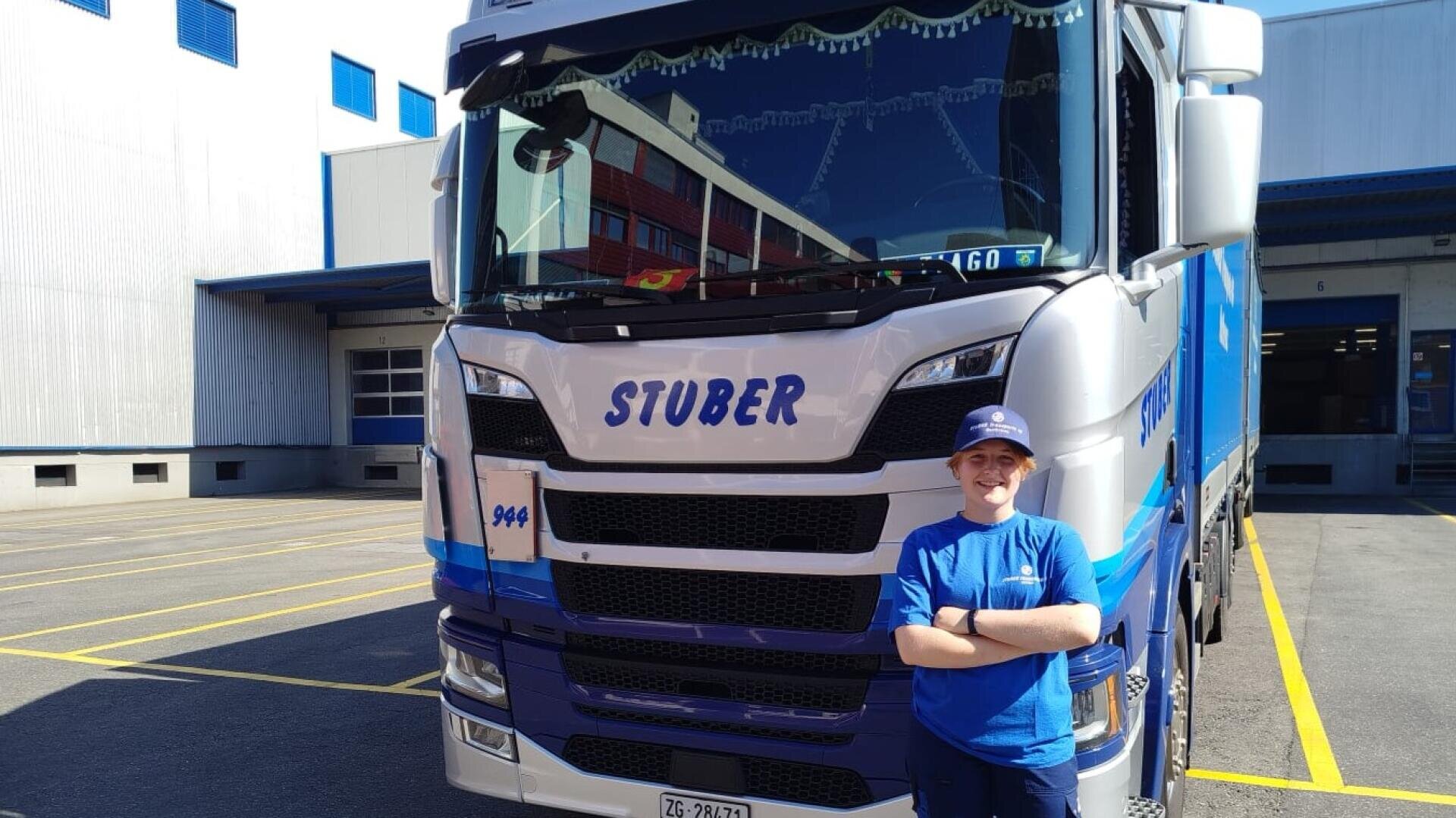 Eine Person in blauer Uniform und Mütze steht lächelnd mit verschränkten Armen vor einem großen weiß-blauen Lastwagen mit der Aufschrift STUBER, der in einem Industriegebiet mit Gebäuden und gelbem Bürgersteig geparkt ist.