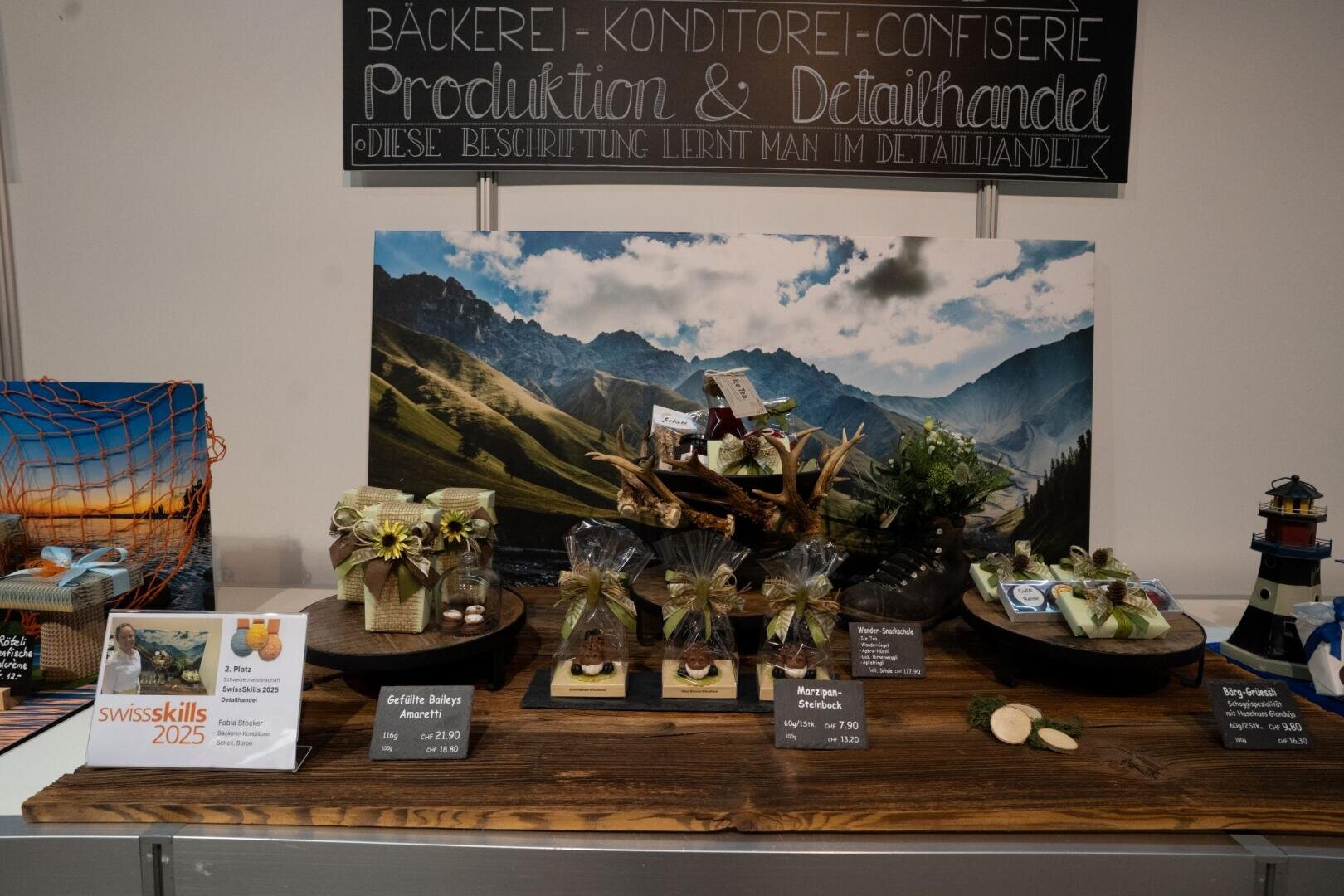 Auf einem Holztisch sind Schweizer Back- und Konditoreiwaren ausgestellt, darunter geschenkverpackte Leckereien, Käse und im Hintergrund ein Foto einer Berglandschaft. Außerdem sind Informationsschilder und Dekorationen zu sehen.