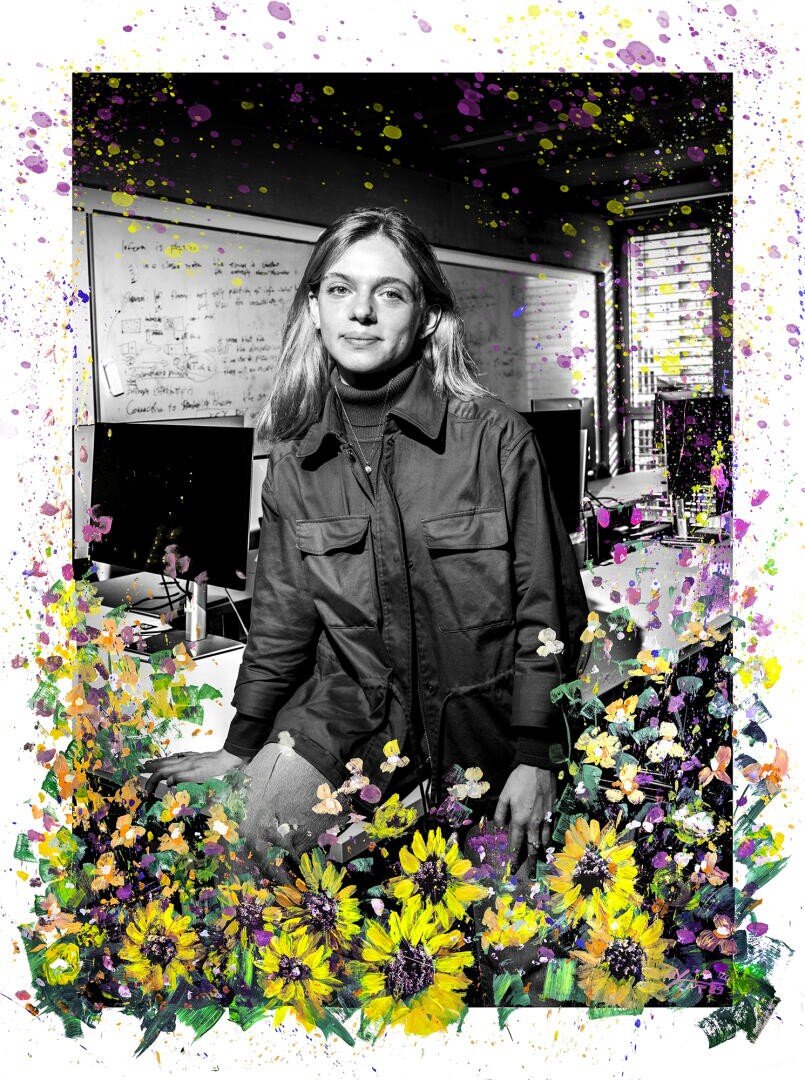 Eine Frau steht in einem Klassenzimmer oder einem Büro, umgeben von Computern und Whiteboards, mit leuchtenden, bunten Spritzern und Blumen, die digital über das Schwarz-Weiß-Foto gelegt werden.