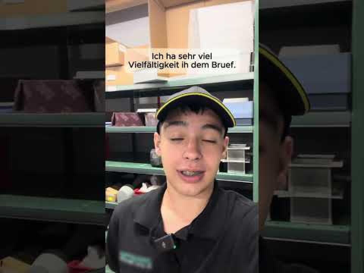 Eine Person in Arbeitsuniform und Mütze steht vor Regalen mit Paketen und Kartons. Der deutsche Text auf dem Bild lautet: "Ich ha sehr viel Vielfalt ih dem Brief.