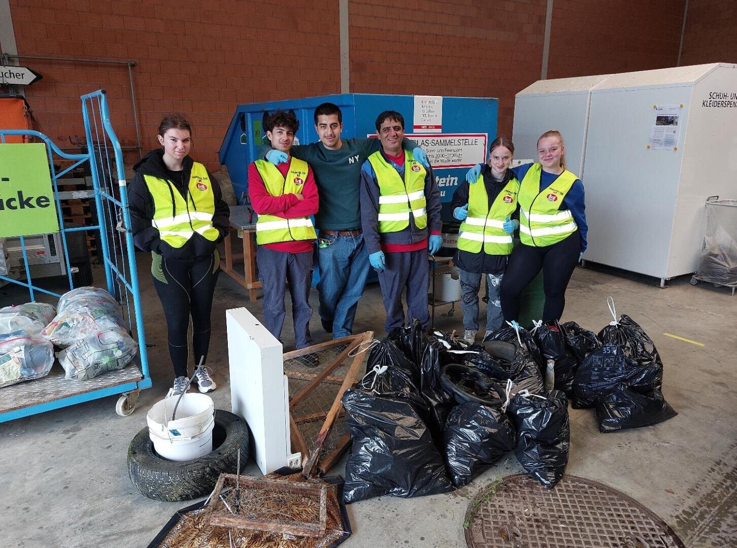 Fünf Personen mit gelben Sicherheitswesten stehen in einem Haus neben einer blauen Recyclingtonne und mehreren gefüllten schwarzen Müllsäcken, Eimern und gesammeltem Abfall; sie scheinen an einer Aufräumaktion teilzunehmen.