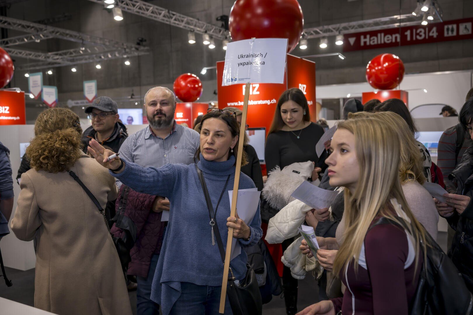 Eine Frau, die ein Schild mit der Aufschrift „Ukrainians/Українці“ in der Hand hält, gestikuliert, während sie von Menschen umringt ist – im Hintergrund sind rote Banner und Stände einer gut besuchten Indoor-Veranstaltung zu sehen.