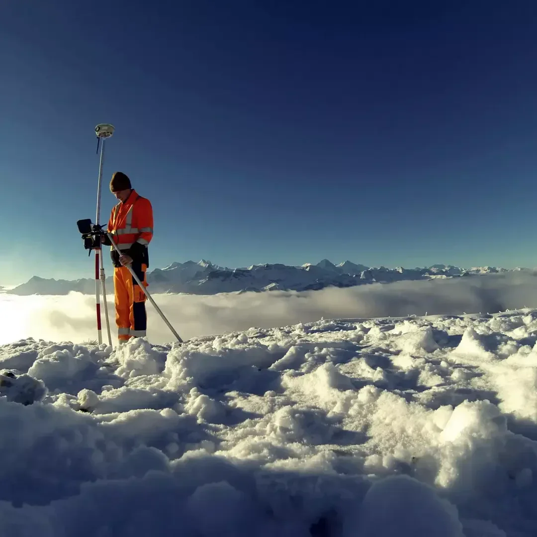 Eine Person in orangefarbener Warnkleidung benutzt eine Vermessungsausrüstung auf einem verschneiten Berggipfel, unter dem Wolken und ferne Berge unter einem klaren blauen Himmel zu sehen sind.