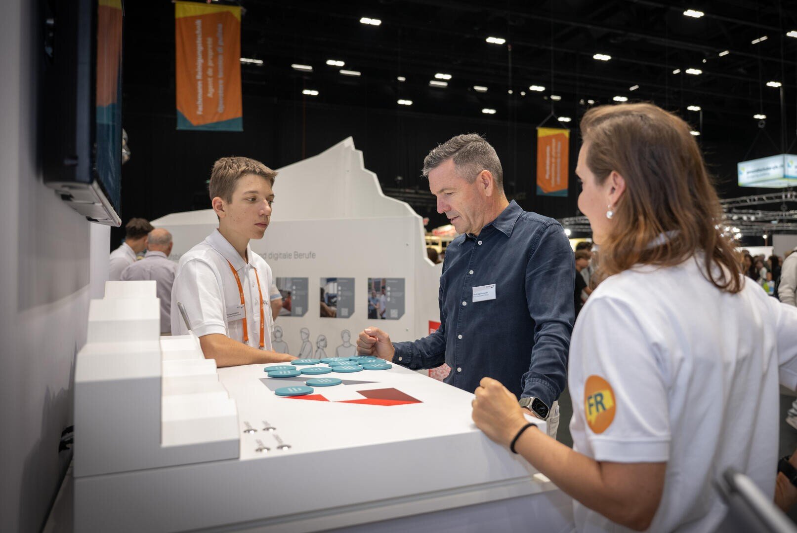 Drei Personen stehen an einem Stand bei einer belebten Veranstaltung in einer Halle und fĂŒhren ein GesprĂ€ch. Zwei tragen weiĂe Hemden mit orangefarbenen Details, die dritte Person ein blaues Hemd. Auf dem Tisch vor ihnen liegen BroschĂŒren und ein weiĂes Display.