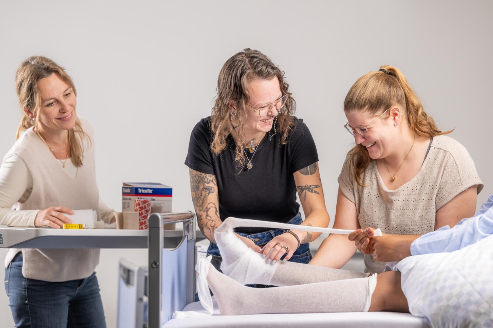 Drei Frauen lächeln und arbeiten zusammen, um in einer hellen klinischen Umgebung ein Bein eines Patienten mit einem medizinischen Verband zu umwickeln. Eine Frau hält das medizinische Material auf einem Wagen, während die anderen den Verband anlegen.