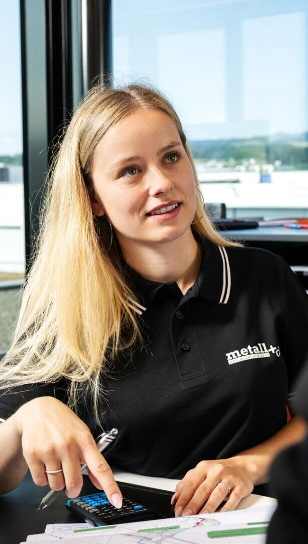 Eine Frau mit langen blonden Haaren, die ein schwarzes Poloshirt aus Metall+ trägt, sitzt an einem Tisch, hält einen Stift und einen Taschenrechner in der Hand, schaut auf und lächelt. Im Hintergrund sind Papiere und ein Fenster mit Blick ins Freie zu sehen.