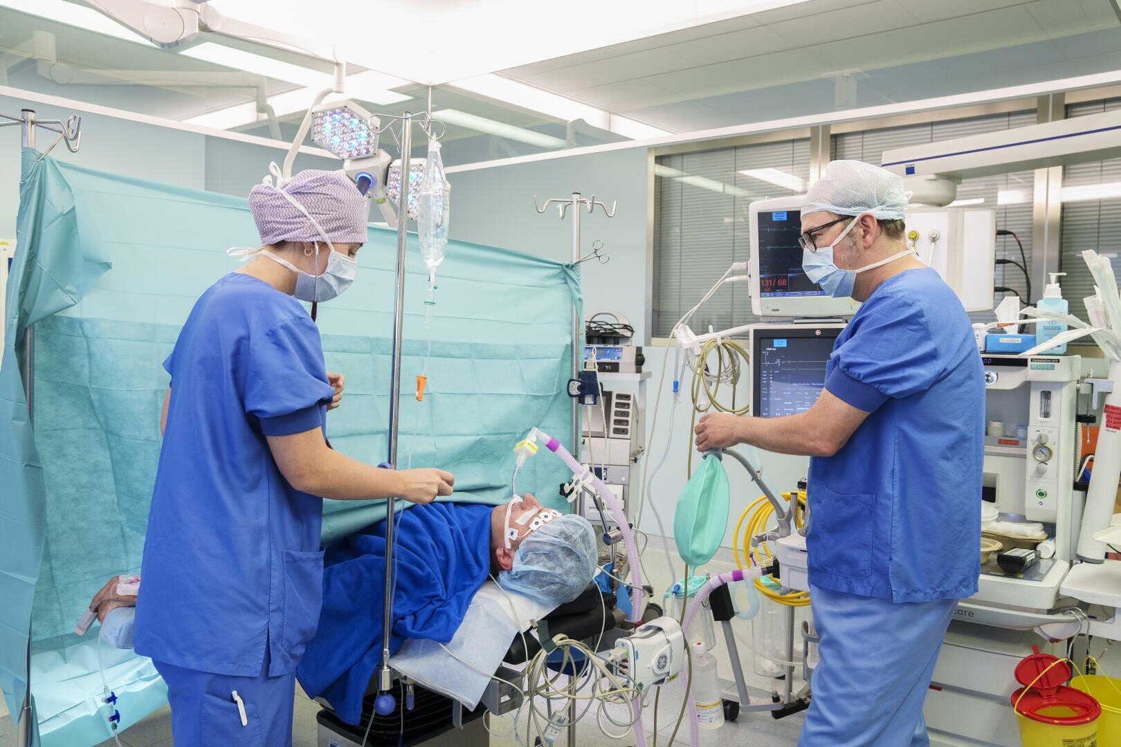 Zwei Mediziner in blauen Kitteln und Masken bereiten einen Patienten in einem Operationssaal auf die Operation vor, wobei sie Geräte und Monitore einstellen und chirurgische Instrumente und Infusionsständer sichtbar sind.
