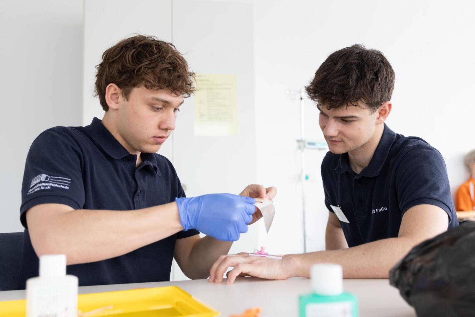 Zwei junge Männer in marineblauen Polohemden sitzen an einem Tisch. Der eine, der blaue Handschuhe trägt, legt dem anderen vorsichtig einen Verband um die Hand und übt einen medizinischen Eingriff. Auf dem Tisch sind medizinische Hilfsmittel und Flaschen zu sehen.