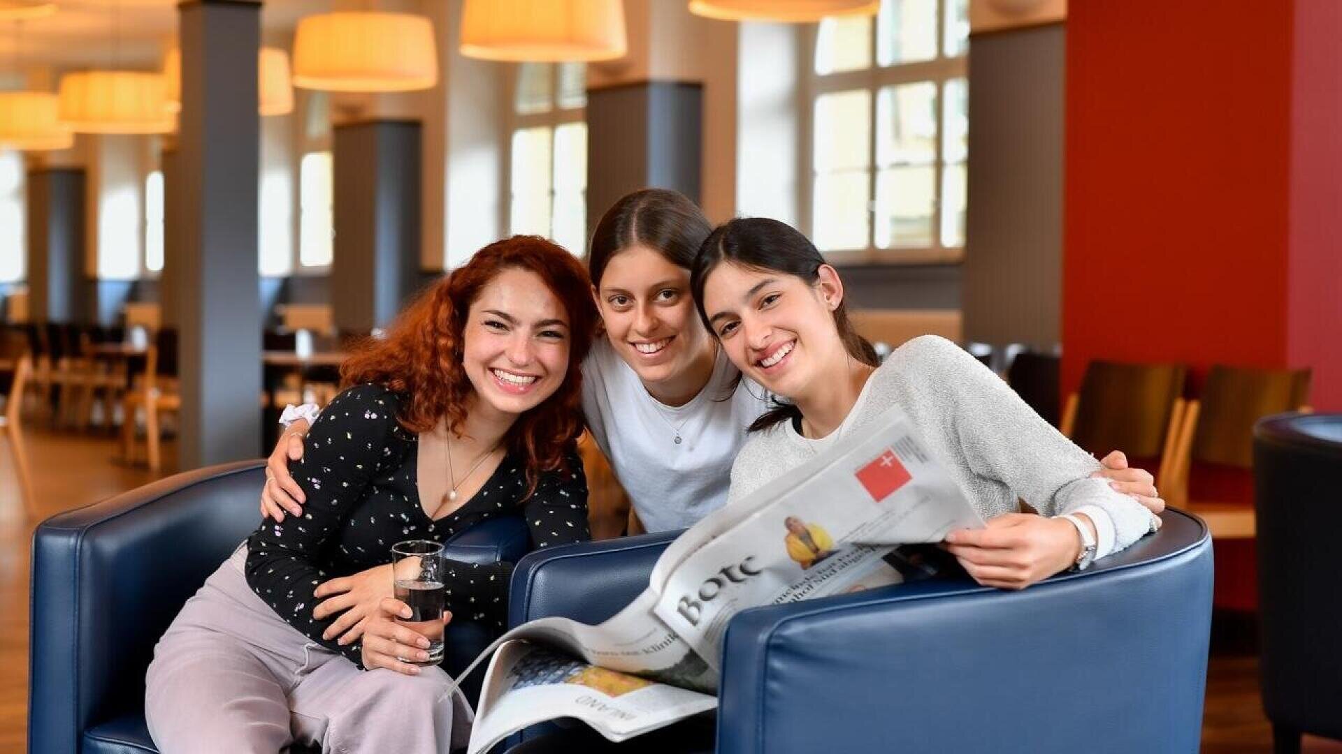 Drei junge Frauen sitzen zusammen in blauen Sesseln; eine von ihnen hält eine Zeitung in der Hand.