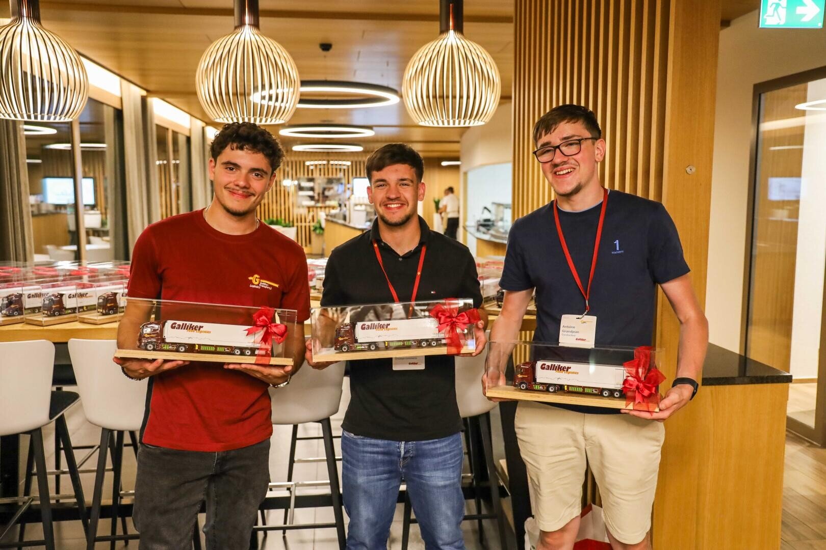 Drei lächelnde junge Männer stehen in einem Raum und halten jeweils eine rechteckige Geschenkbox mit einer roten Schleife in der Hand. Sie tragen Umhängebänder und Freizeitkleidung und befinden sich in einer modernen Umgebung mit Holzdekor und Hängeleuchten.