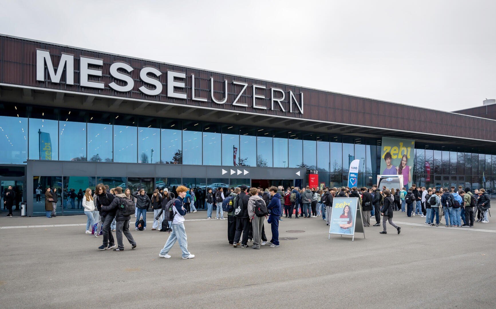An einem bewölkten Tag versammeln sich Menschenmassen vor dem gläsernen Kongresszentrum der Messe Luzern. Einige stehen in Gruppen, während andere in der Nähe des Eingangs Schlange stehen. Veranstaltungsschilder und Transparente sind sichtbar.