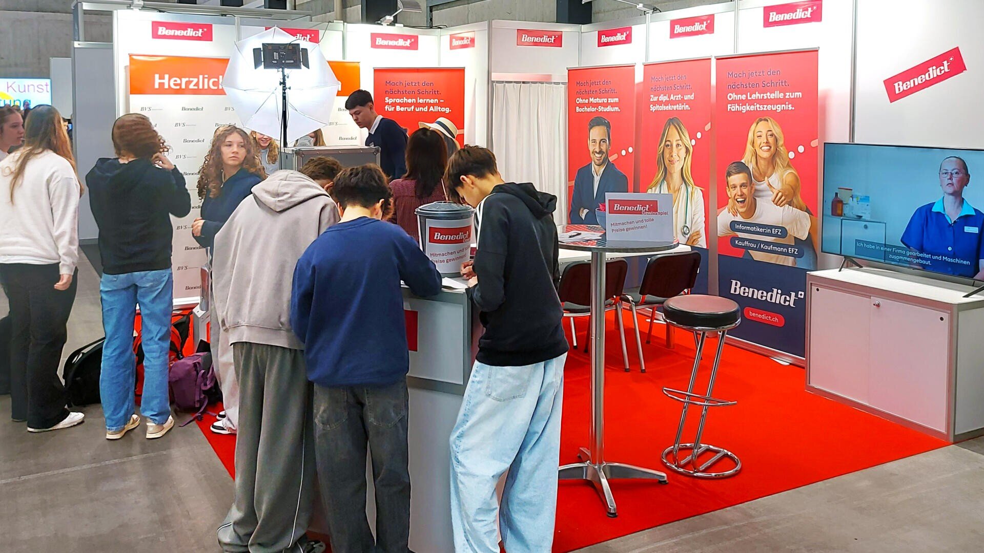 Eine Gruppe von Schülern versammelt sich an einem Informationsstand der Benedict-Schule auf einer Berufsmesse, an dem rote Banner mit dem Schullogo und ein Fernsehbildschirm mit Unternehmensinformationen zu sehen sind.