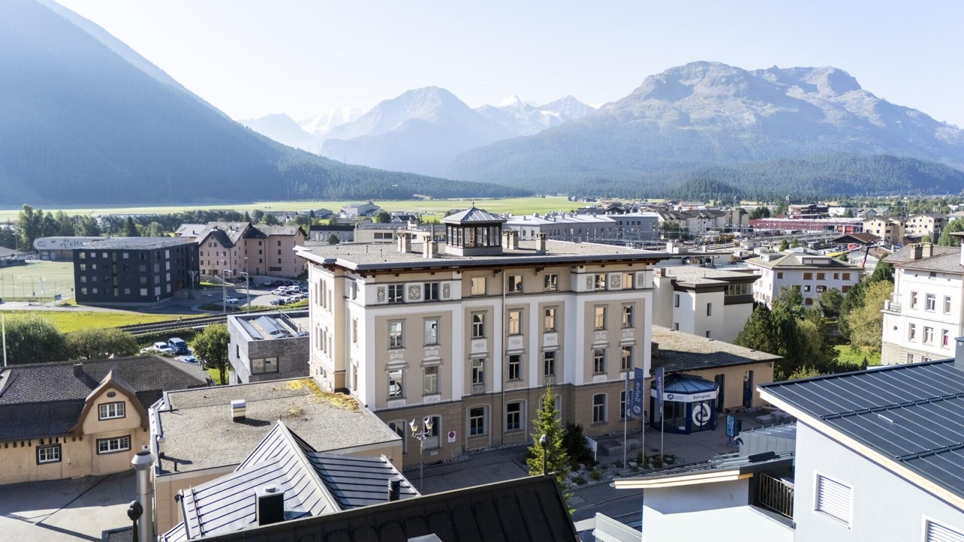 Luftaufnahme von Samedan mit einem Teil der Campus der Höheren Fachschule für Tourismus & Management in Graubünden, gelegen im Engadin, umgeben von grünen Feldern und hohen Bergen unter einem strahlend blauen Himmel.