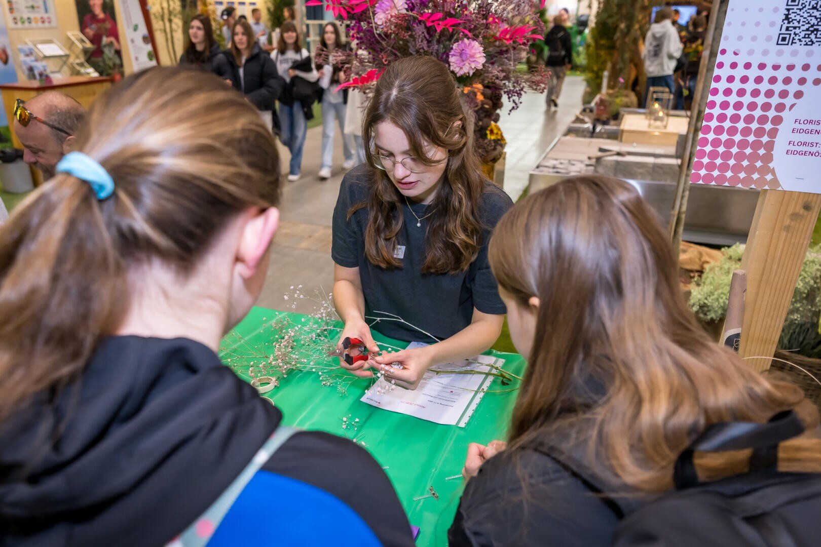 Eine Frau zeigt zwei jungen Leuten an einem Tisch mit floralen Materialien, wie man Blumen arrangiert, während im Hintergrund andere Leute bei einer Indoor-Veranstaltung vorbeigehen.