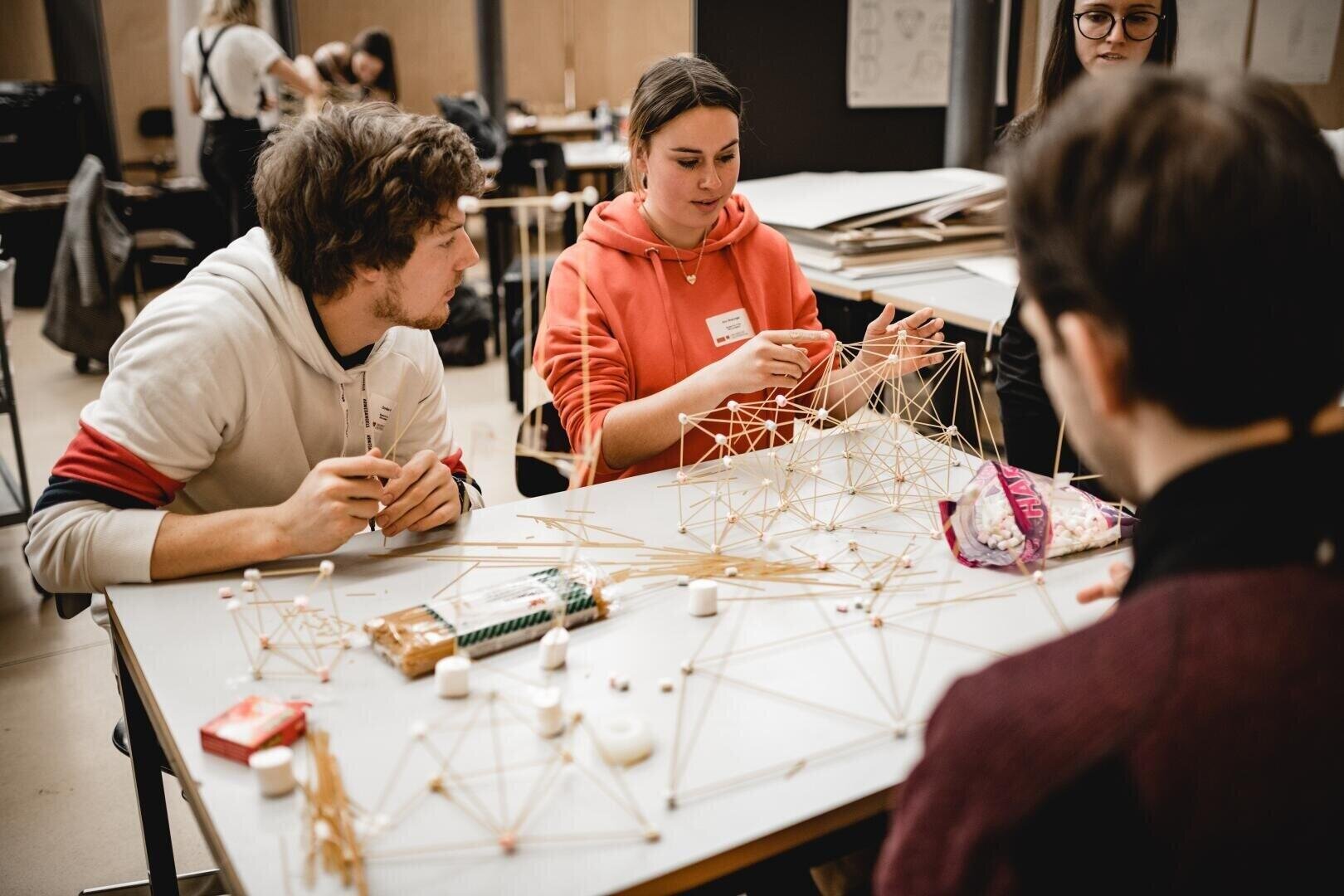 Drei junge Erwachsene sitzen an einem Tisch und bauen geometrische Strukturen aus Spaghetti und Marshmallows. Sie arbeiten gemeinsam an einem Projekt in einer lockeren Schulumgebung. Das Material und die fertigen Modelle liegen verstreut auf dem Tisch.