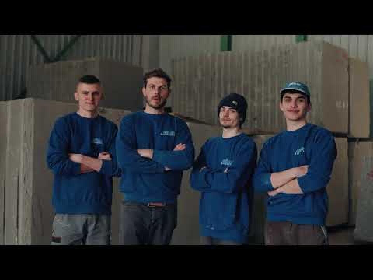 Vier junge Männer in passenden blauen Sweatshirts stehen mit verschränkten Armen in einer Lagerhalle und posieren gemeinsam vor gestapelten Kartons oder Paletten.