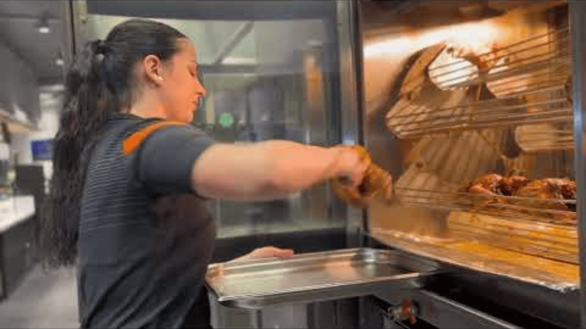 Eine Frau nimmt in einer Großküche gebratene Hähnchen mit einem Tablett aus einem Rotisserie-Ofen heraus.