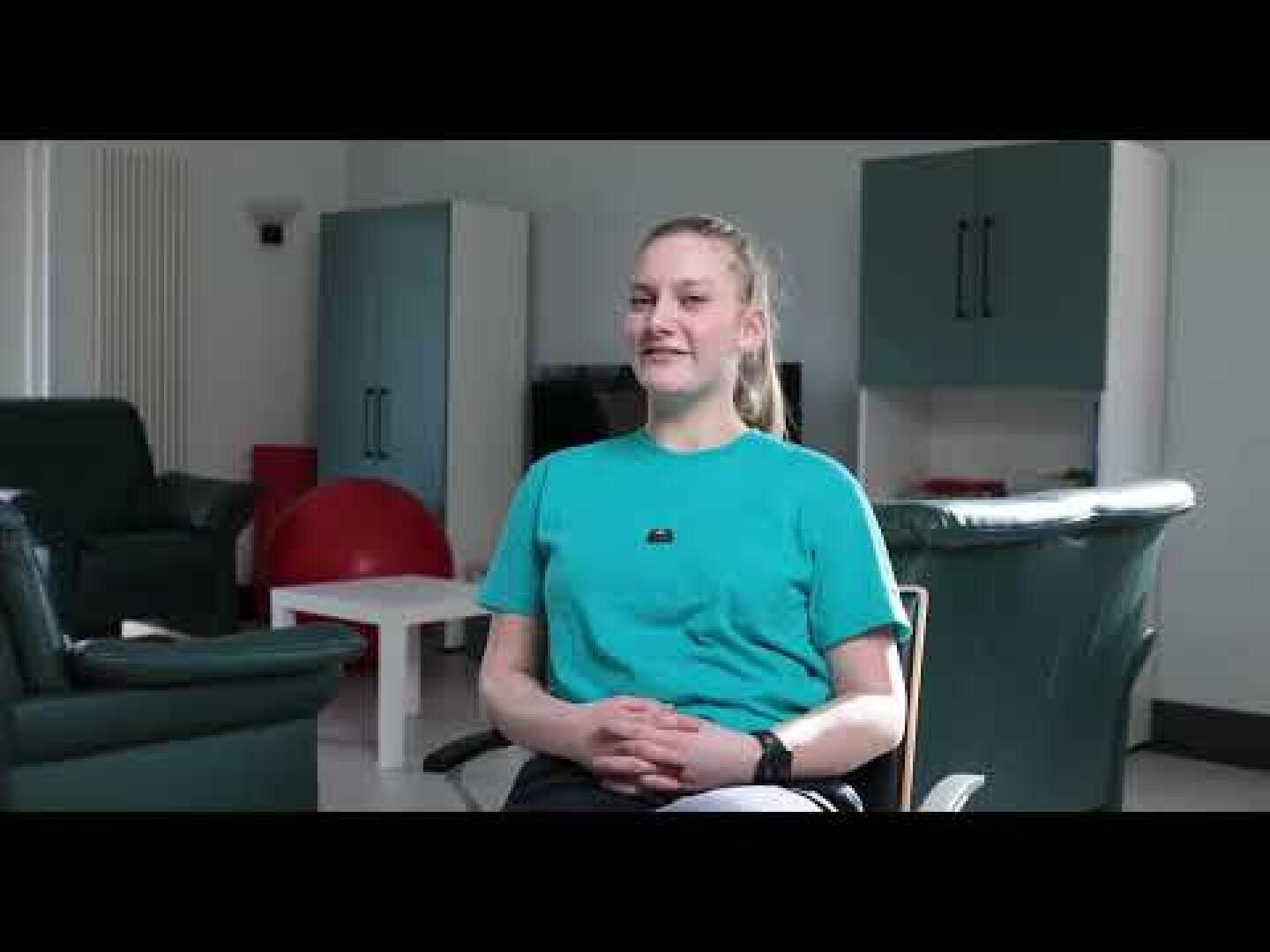 Eine Person sitzt in einem Wohnzimmer mit grünen Stühlen, Schränken und einem roten Gymnastikball im Hintergrund und lächelt.