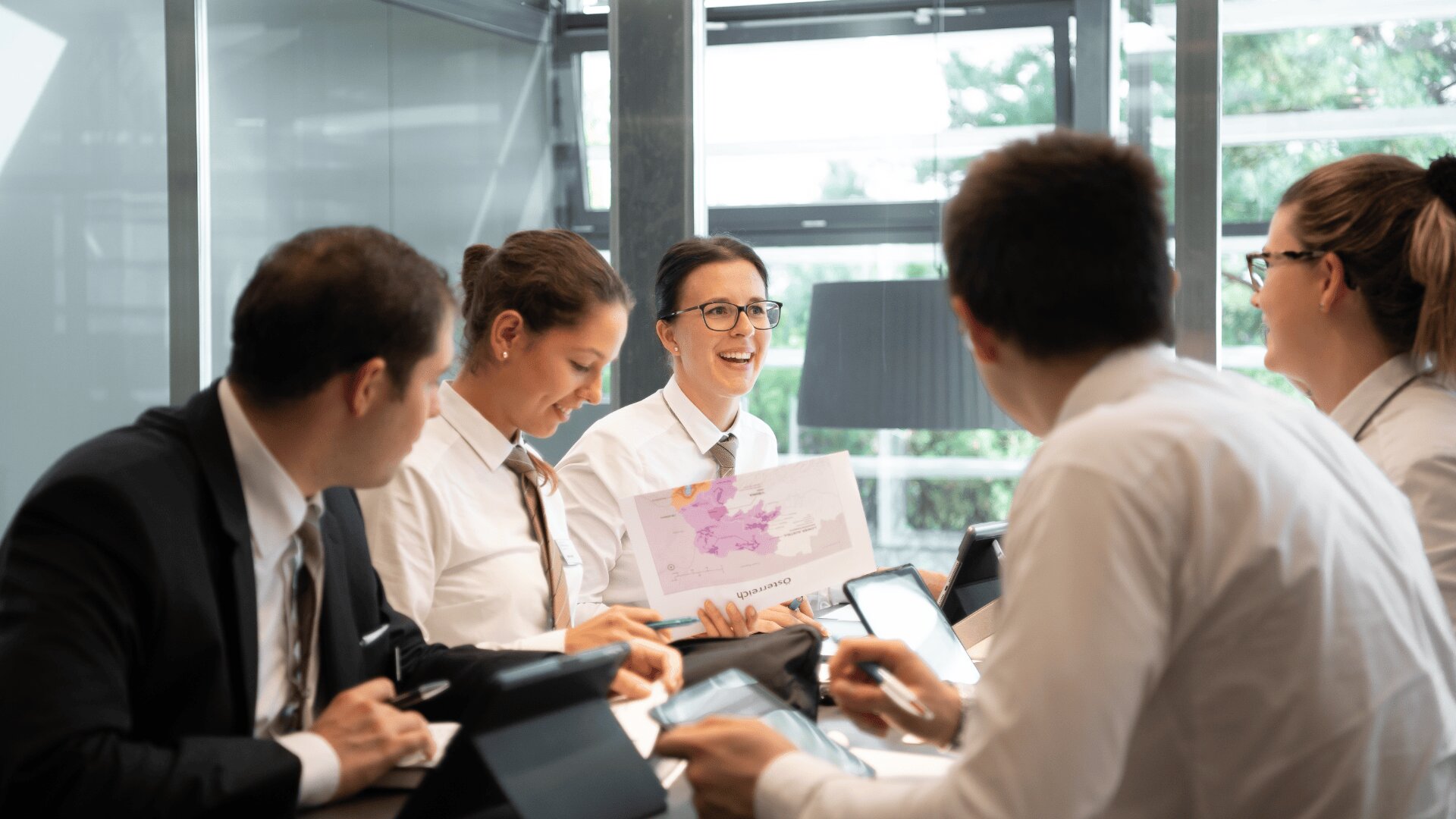 Fünf Personen in weißen Hemden und Krawatten sitzen um einen Konferenztisch und diskutieren. Eine Person lächelt und hält eine Karte oder ein Diagramm hoch. Auf dem Tisch sind Laptops und Papiere zu sehen, mit großen Fenstern im Hintergrund.