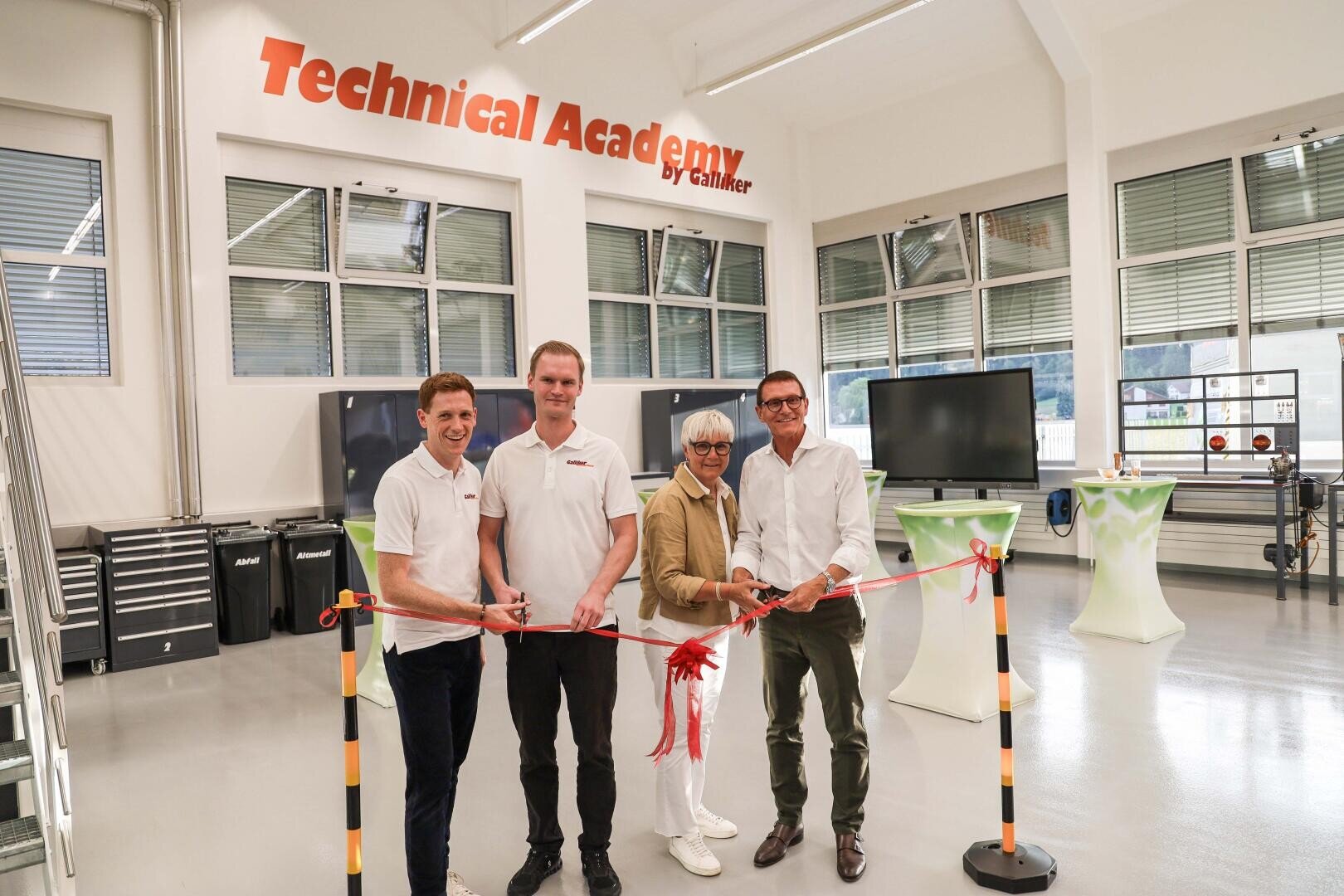 Vier Personen stehen in einem hellen Klassenzimmer mit der Aufschrift „Technical Academy“ und halten ein rotes Band für eine Feier in den Händen. Zwei tragen weiße Hemden, eine Person eine helle Jacke und eine Brille, und eine weitere Person ein weißes Hemd und eine dunkle Hose.