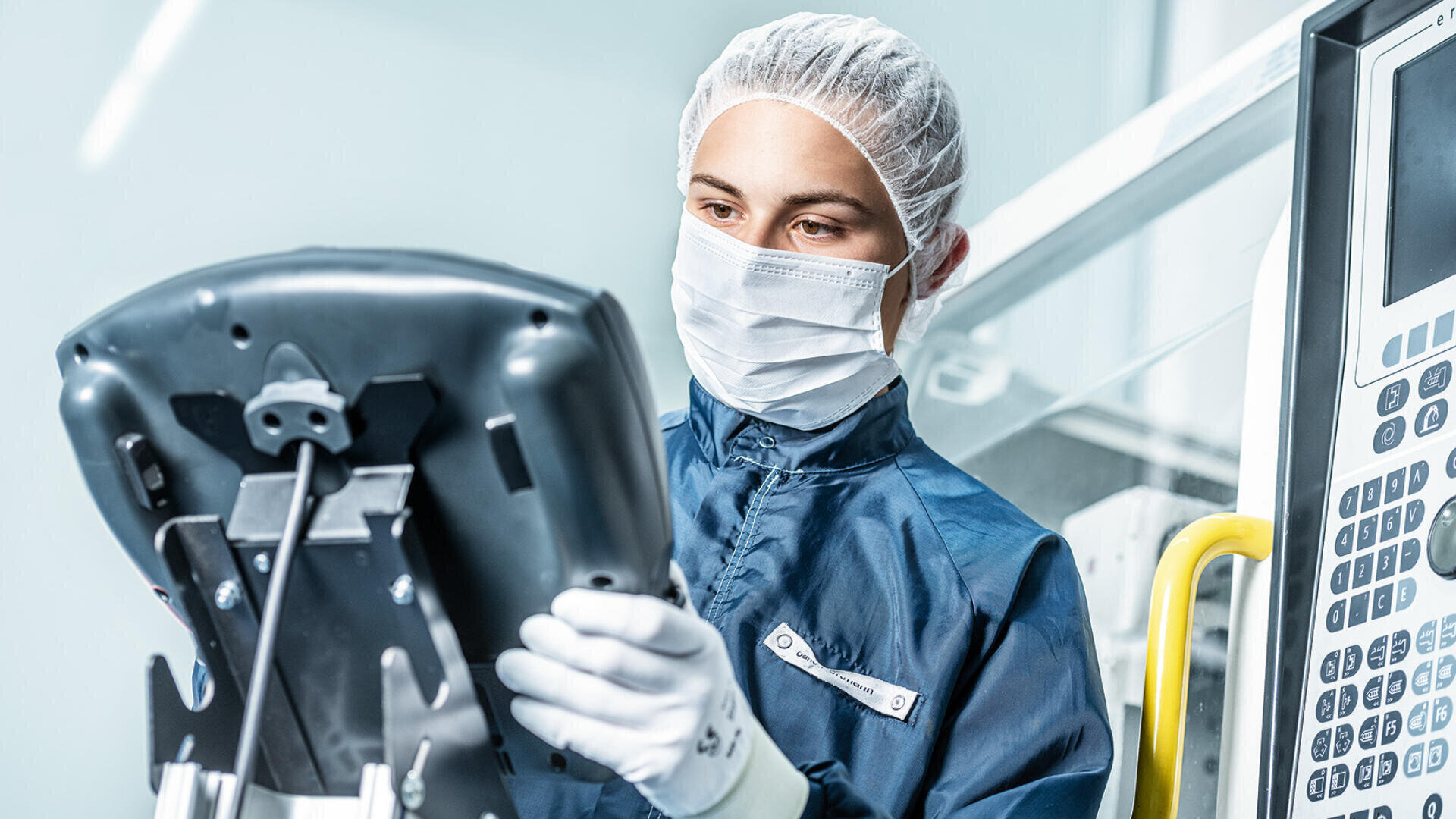 Eine Person, die Schutzkleidung, eine Gesichtsmaske, Handschuhe und ein Haarnetz trägt, bedient ein Touchscreen-Gerät in einem sauberen Hightech-Labor oder einer Produktionsumgebung.