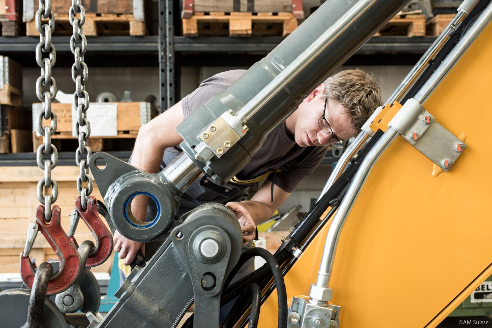 Eine Person mit Schutzbrille untersucht und arbeitet an einer großen Industriemaschine oder einem hydraulischen Arm in einer Werkstatt mit Regalen und Werkzeugen im Hintergrund.