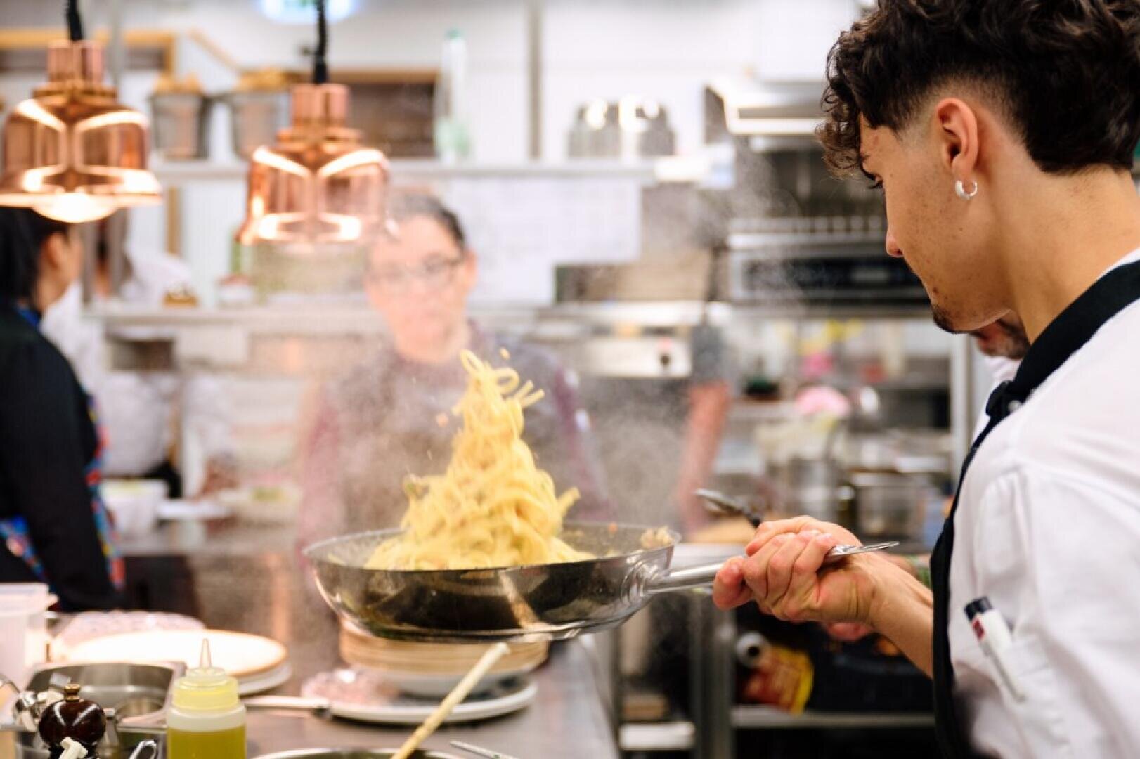 Ein Koch schwenkt Nudeln in einer Pfanne, wobei Dampf in einer geschäftigen Restaurantküche aufsteigt, während im Hintergrund andere Mitarbeiter zu sehen sind.