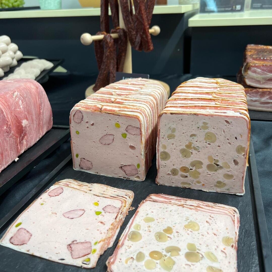 Rechteckige Blöcke mit in Scheiben geschnittenem Fleisch, teilweise mit eingebetteten Pistazien und anderen Zutaten, werden auf einer Theke neben anderen Wurstsorten ausgestellt.