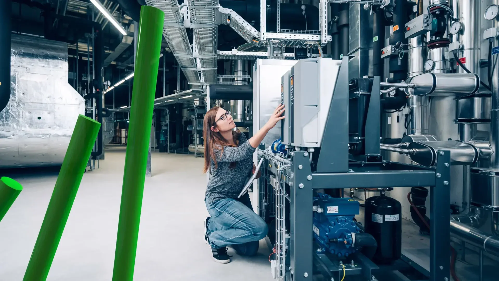 Eine Frau mit Brille und Freizeitkleidung kniet in einer modernen, geräumigen Fabrikumgebung mit freiliegenden Rohren und Metallstrukturen und stellt die Bedienelemente von Industriemaschinen ein.