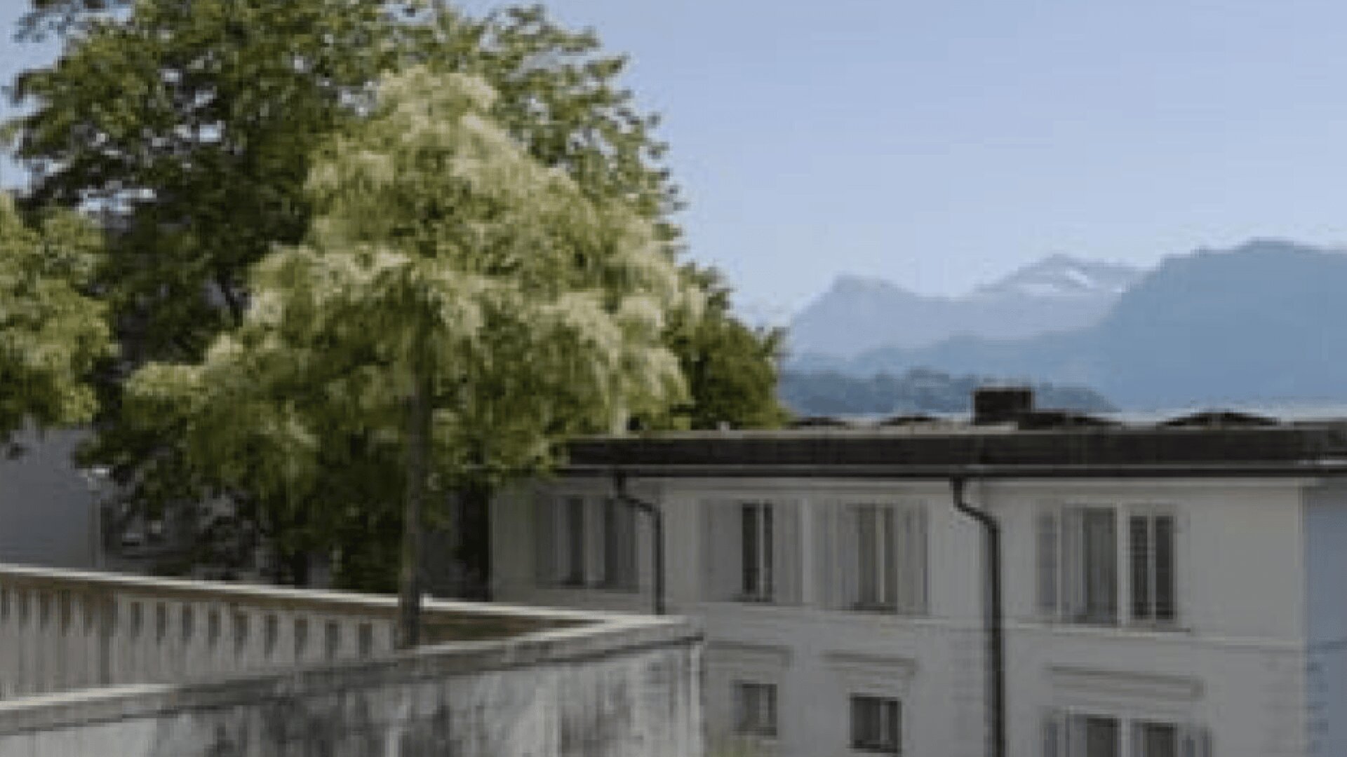 Ein belaubter Baum steht neben einem hellen Gebäude mit weiß umrahmten Fenstern vor der Kulisse der Berge unter einem klaren Himmel.