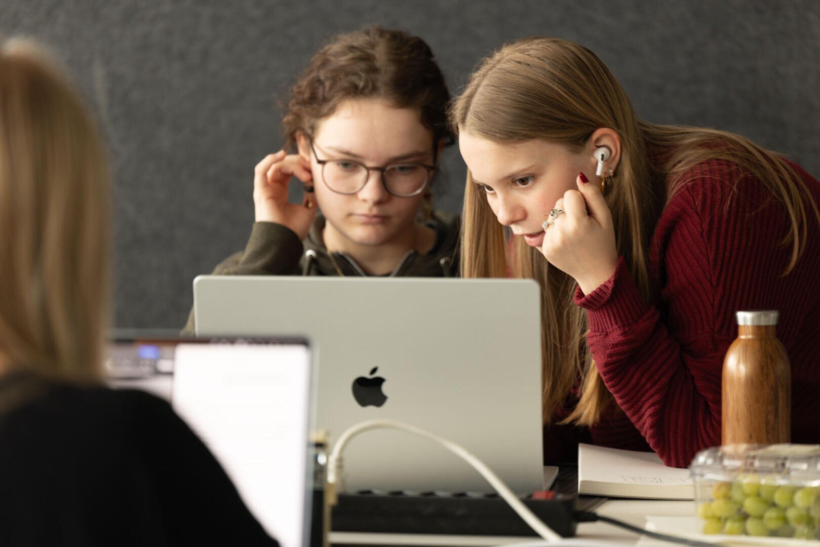 Zwei Mädchen konzentrieren sich auf einen Laptop, eine mit Brille, die andere mit Kopfhörern. Sie sitzen an einem Tisch mit Notizbüchern, einer Wasserflasche und einem Behälter mit Weintrauben, während eine andere Person in der Nähe an einem Laptop arbeitet.