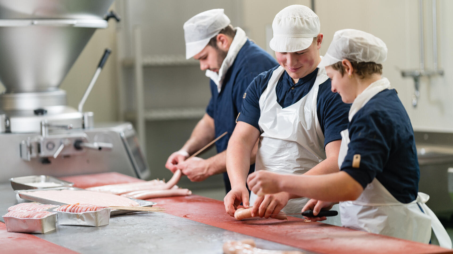 Drei Arbeiter mit weißen Schürzen und Hüten bereiten in einem Lebensmittelverarbeitungsbetrieb gemeinsam Würste zu, wobei sie Messer benutzen und die Würste auf Tabletts auf einer langen roten Arbeitsfläche ablegen.