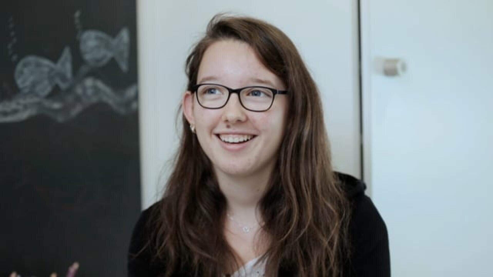 Eine junge Frau mit langen braunen Haaren und Brille sitzt lächelnd in einem Raum. Hinter ihr befindet sich eine Kreidetafel mit Fischzeichnungen und eine weiße Wand.