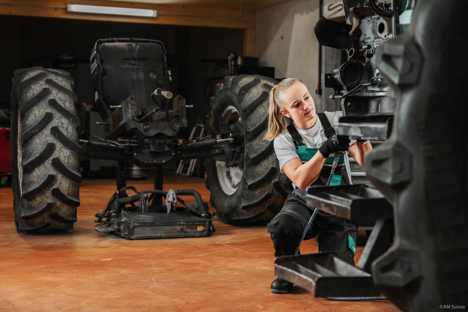 Eine Frau in Arbeitsoverall und Handschuhen kniet in einer Werkstatt neben einem großen Traktorreifen und benutzt ein Werkzeug, um Schrauben am Rad anzuziehen oder zu lösen. Im Hintergrund ist ein weiterer Traktorrahmen zu sehen.