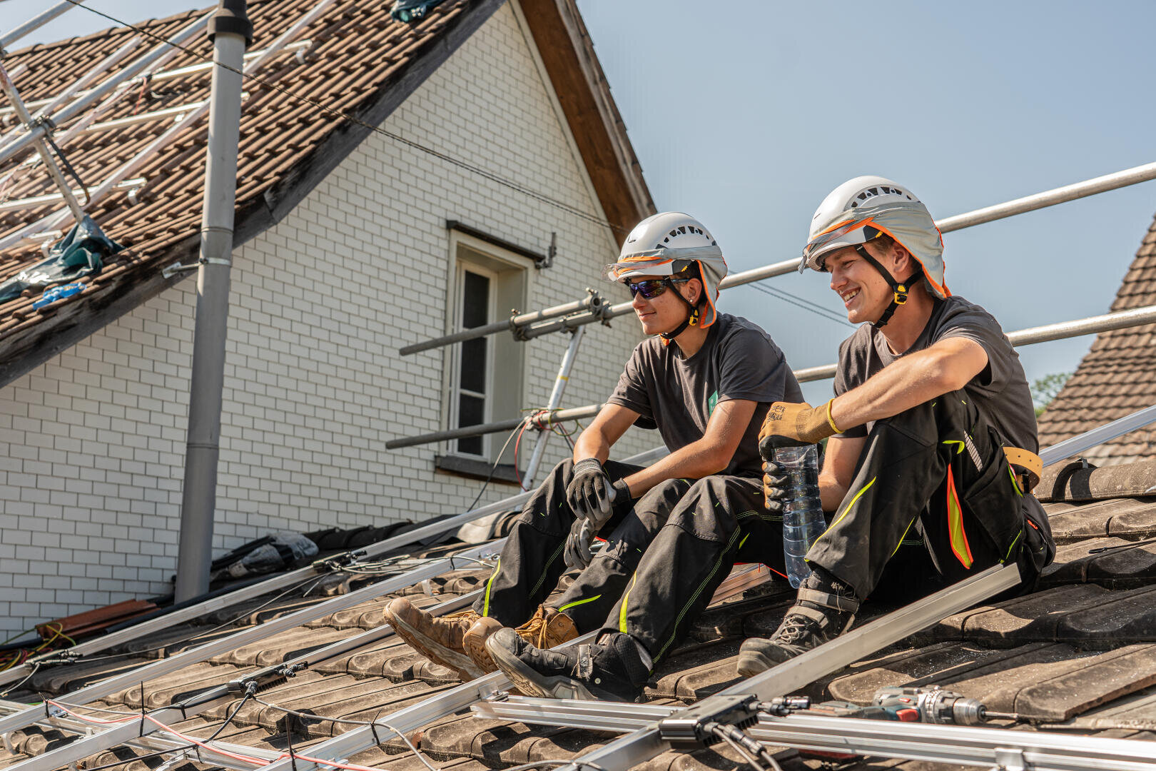 Zwei Bauarbeiter mit Helmen und Sicherheitsausrüstung sitzen auf einem Hausdach, lächeln und machen eine Pause inmitten von Gerüsten und Geräten für die Installation von Solarmodulen unter einem klaren Himmel.