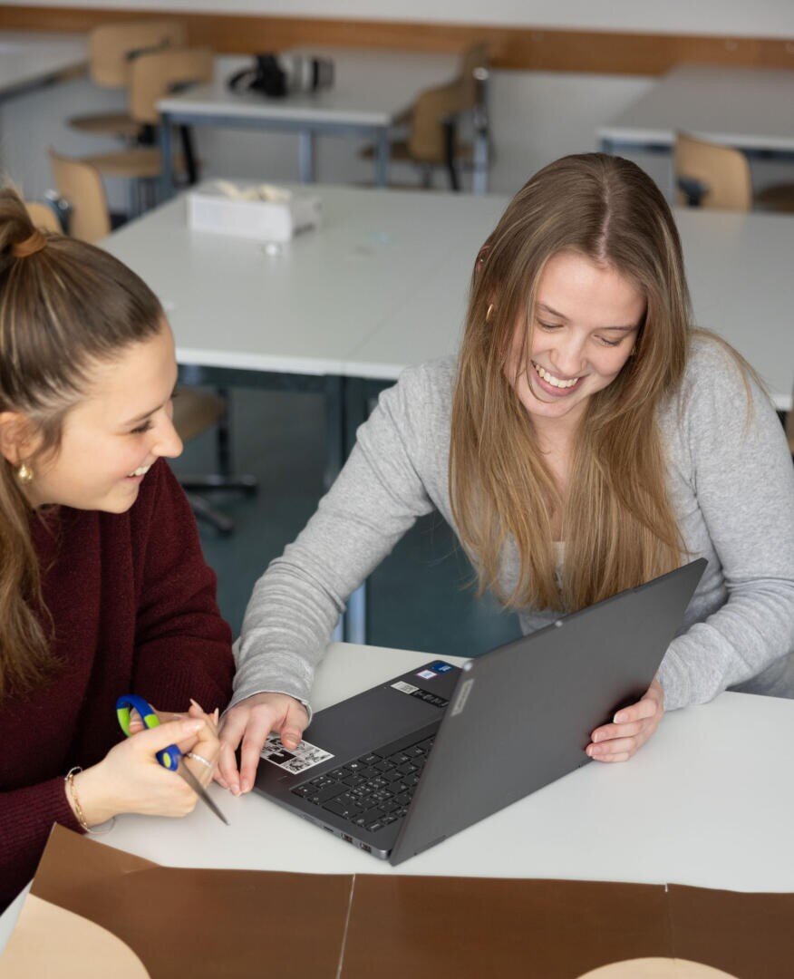 Zwei junge Frauen sitzen an einem Schreibtisch in einem Klassenzimmer, lächeln und schauen gemeinsam auf einen Laptop-Bildschirm. Eine hält eine Schere, und auf dem Tisch liegt Papier. Im Hintergrund sind leere Tische und Stühle zu sehen.
