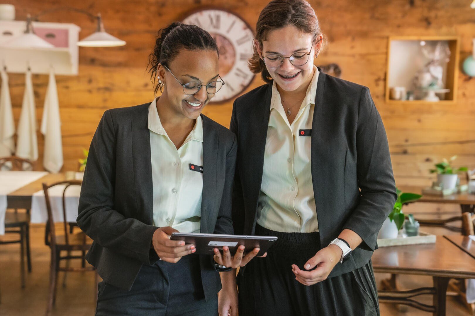 Zwei Frauen in Geschäftskleidung stehen zusammen in einem warm beleuchteten Restaurant und schauen lächelnd auf eine Tafel. Die Kulisse besteht aus Holzwänden, einer Uhr und Topfpflanzen im Hintergrund.