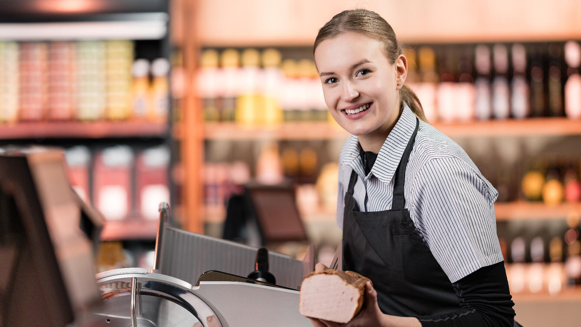Eine lächelnde Frau in einer Schürze hält ein Stück Feinkostfleisch hinter einer Theke, im Hintergrund sind Regale mit Flaschen und Produkten zu sehen.