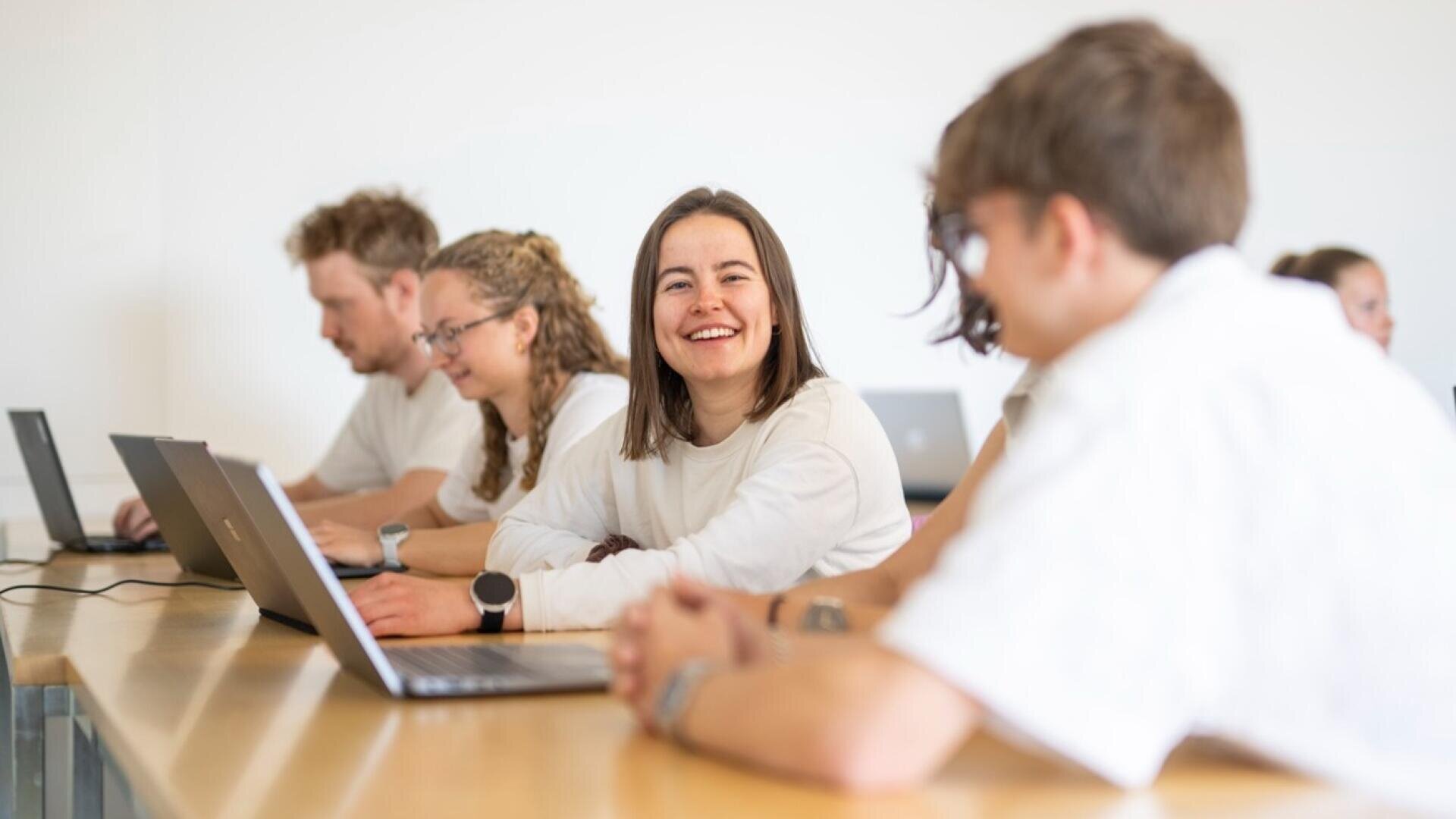 Eine Gruppe von Menschen sitzt an einem langen Tisch mit Laptops und unterhält sich. Eine Frau in der Mitte, die lächelt und in die Kamera schaut, ist umgeben von anderen, die in einem hellen, weißwandigen Raum arbeiten und sich unterhalten.