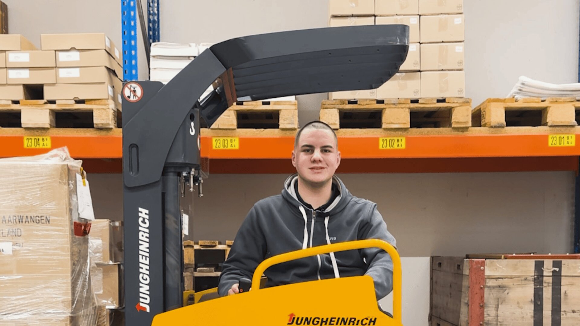 Ein Mann in einem grauen Kapuzenpulli bedient einen Jungheinrich-Gabelstapler in einem Lagerhaus, umgeben von Regalen mit gestapelten Kisten und Holzpaletten.