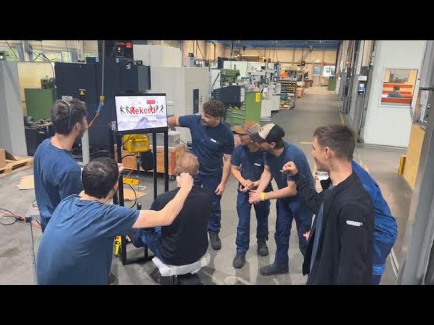 Eine Gruppe von acht Personen in Arbeitsuniformen versammelt sich um einen Bildschirm in einer Industriehalle und betrachtet eine Folie mit der Aufschrift MekoBot2! Im Hintergrund sind Maschinen und Arbeitsstationen zu sehen.
