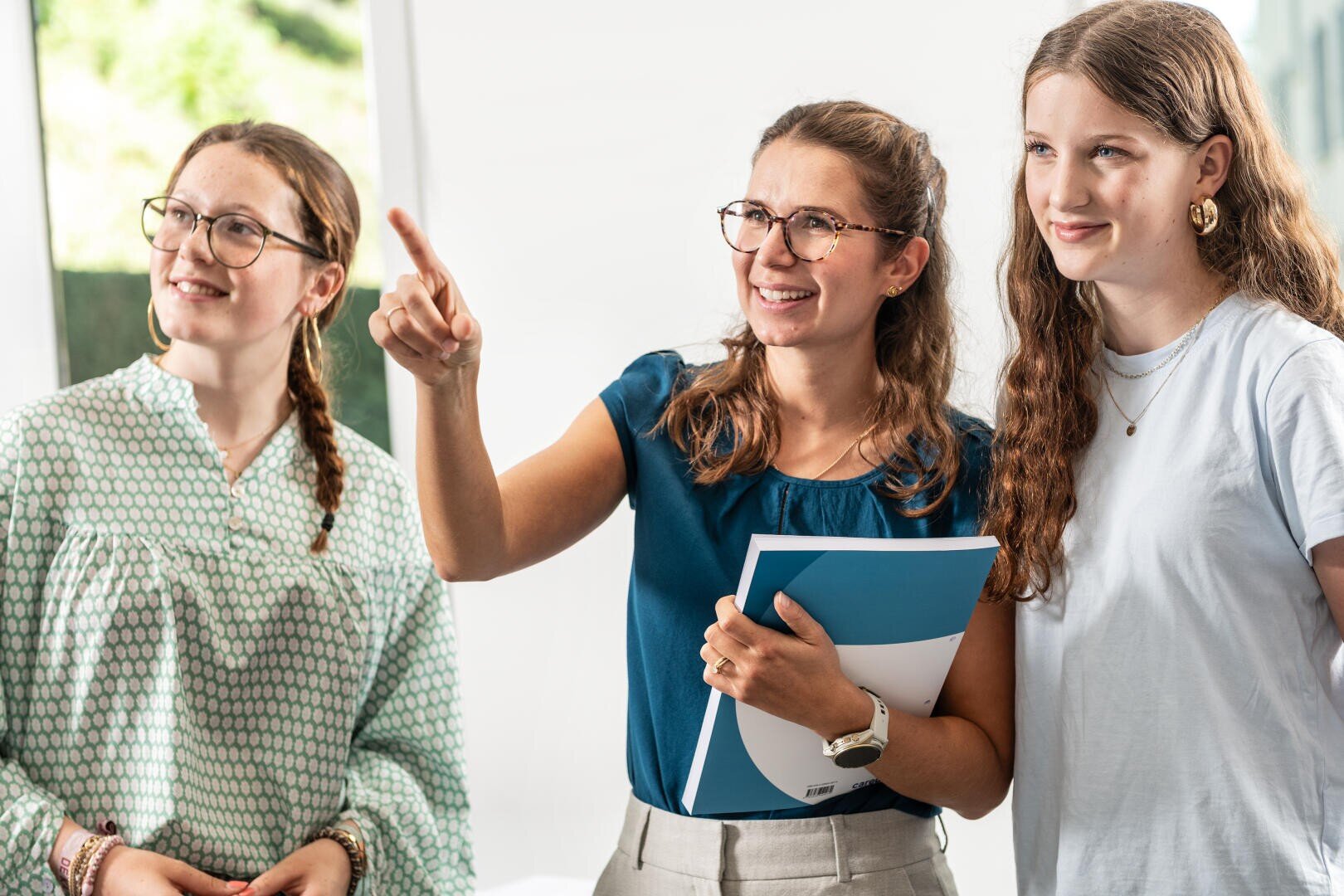 Drei junge Frauen stehen in einem Innenraum zusammen. Eine Frau in der Mitte, die Dokumente in der Hand hält, lächelt und deutet nach vorne, während die beiden anderen in dieselbe Richtung schauen und alle engagiert und fröhlich wirken.