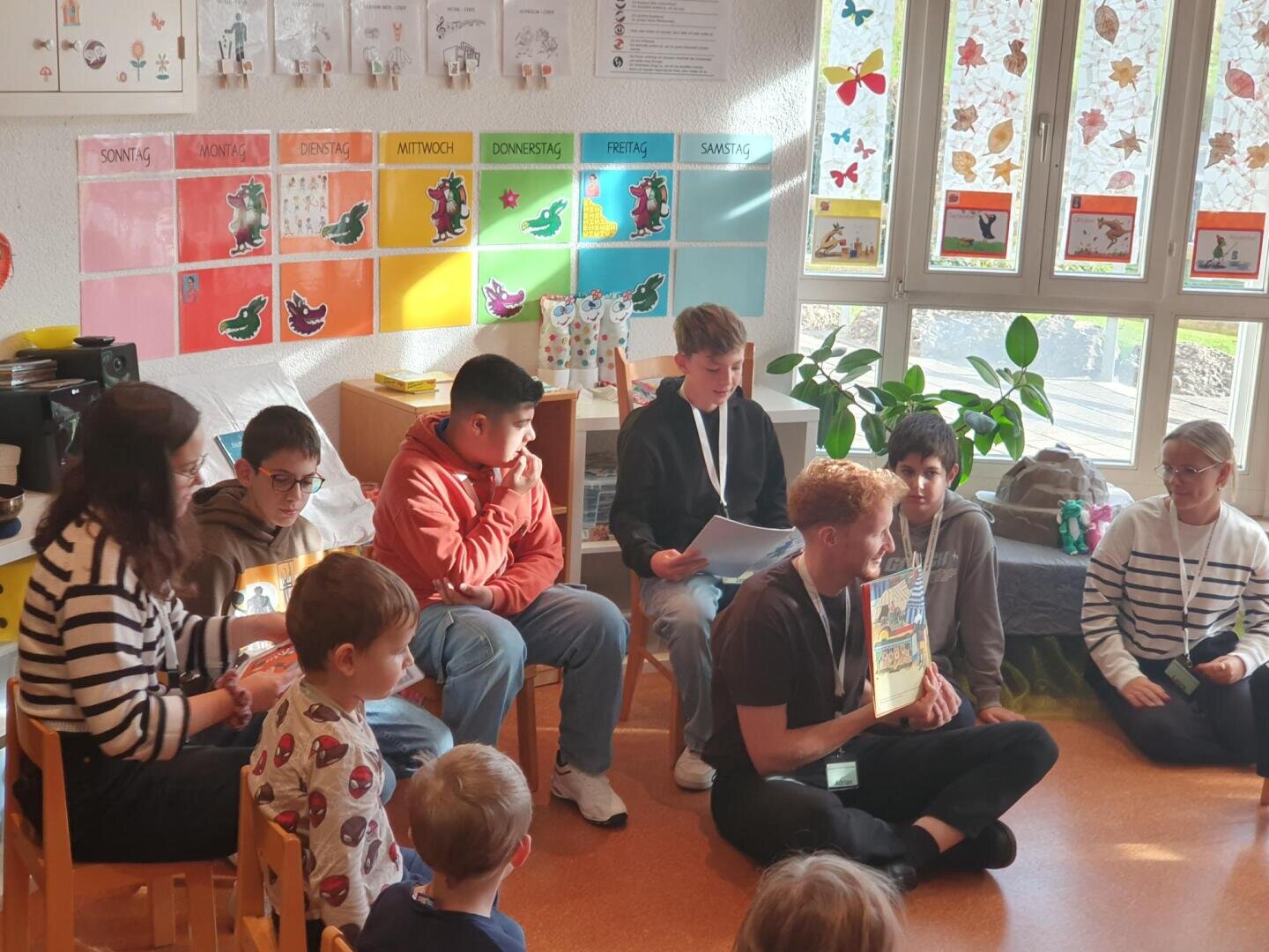 Eine Gruppe von Kindern und Jugendlichen sitzt in einem farbenfrohen Klassenzimmer, während eine Person aus einem Buch vorliest. An der Wand hängen bunte Zeichnungen und Karten mit den Wochentagen; Sonnenlicht strömt durch die verzierten Fenster herein.