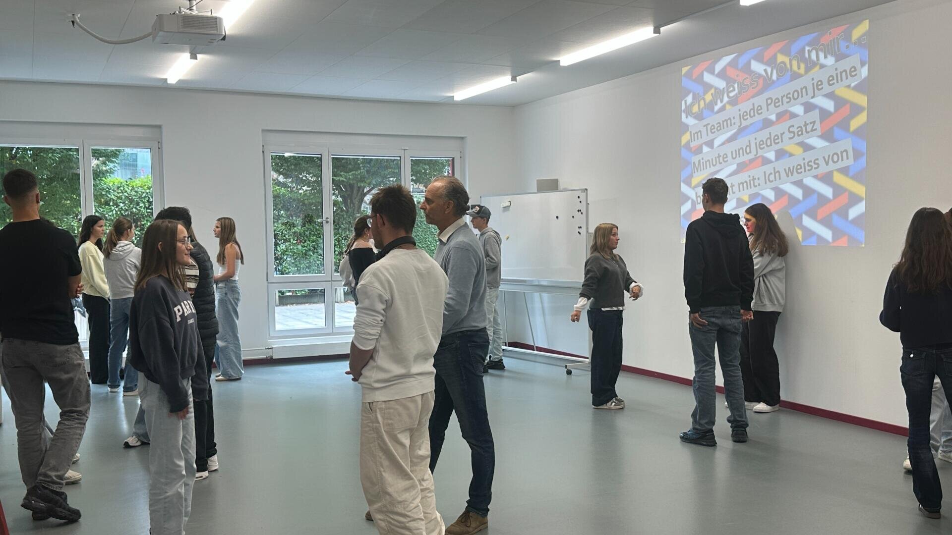 Eine Gruppe von Menschen steht in Paaren und kleinen Gruppen in einem hellen Klassenzimmer und unterhält sich. Eine bunte Folie mit deutschem Text wird an die Wand projiziert. Große Fenster im Hintergrund zeigen Grünflächen im Freien.