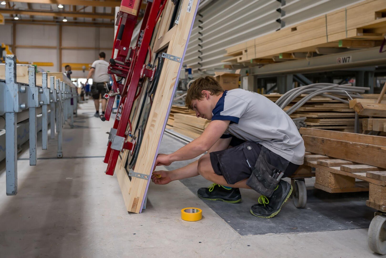 Ein Junge in Arbeitskleidung kniet auf dem Boden einer Werkstatt und misst oder justiert einen großen Holzrahmen mit Werkzeugen in der Nähe. Im Hintergrund sind Holzstapel und verschiedene Geräte zu sehen.