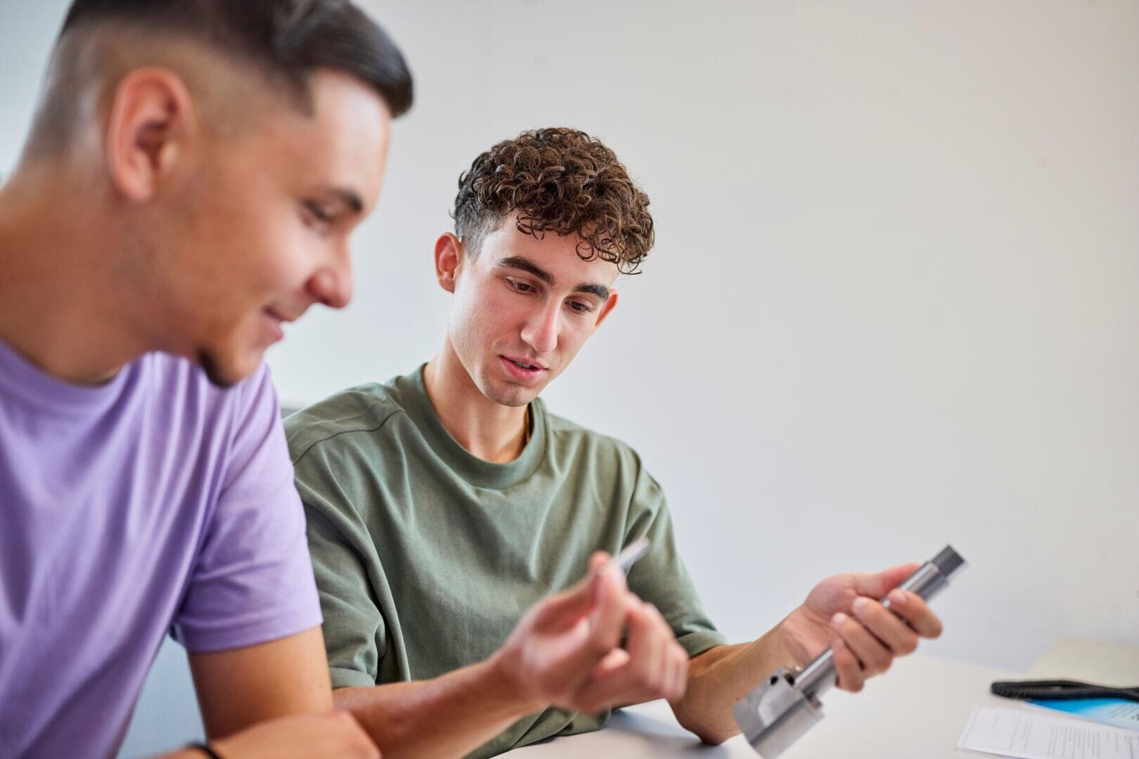 Zwei junge Männer sitzen an einem Tisch; einer hält einen Metallgegenstand in der Hand und erklärt dem anderen etwas. Beide wirken konzentriert und sind in ein Gespräch vertieft; im Hintergrund sind Papiere und eine neutrale Kulisse zu sehen.