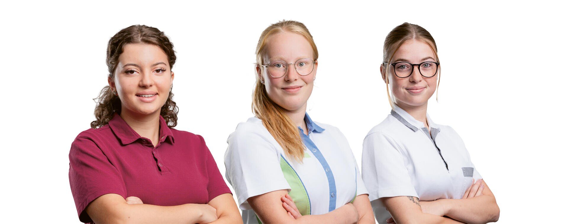 Drei junge Frauen stehen Seite an Seite, die Arme verschränkt, und lächeln. Vor einem weißen Hintergrund tragen sie Arbeitsuniformen in verschiedenen Farben.