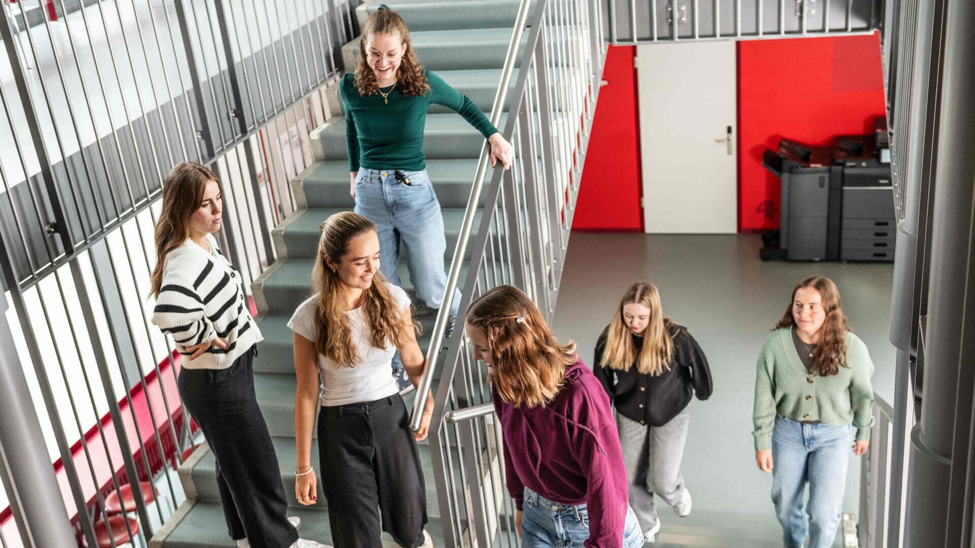 Sechs junge Frauen gehen und unterhalten sich gemeinsam auf einer Treppe in einem modernen Gebäude mit grauem Geländer, einer rot-weißen Wand und Büroausstattung im Hintergrund. Sie scheinen Studentinnen oder Freundinnen zu sein.