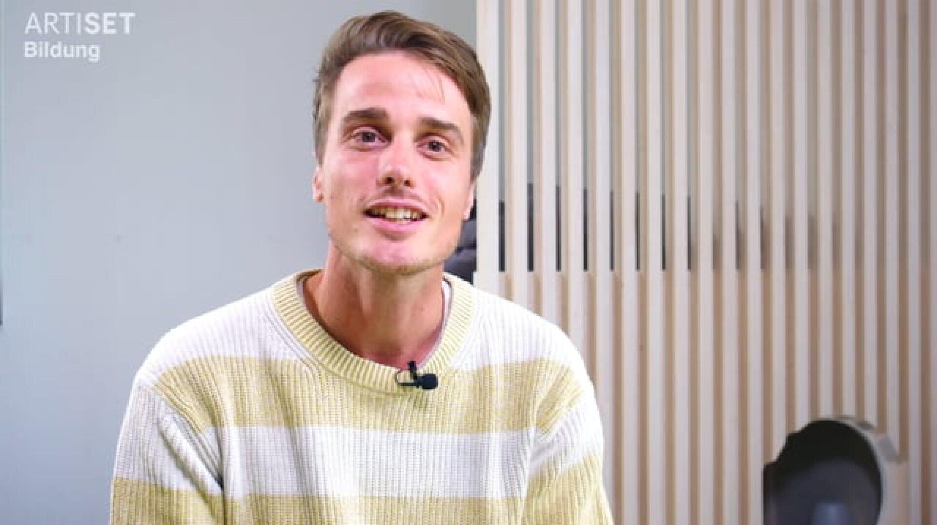 Ein Mann mit kurzen braunen Haaren, der einen weiß-gelb gestreiften Pullover trägt, sitzt in einem Raum und lächelt in die Kamera. An seinem Pullover ist ein Mikrofon befestigt. Im Hintergrund sind vertikale Holzlatten und eine graue Wand zu sehen.